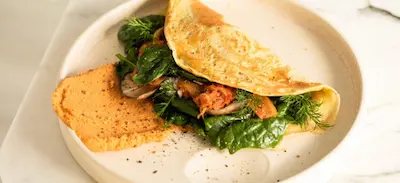 Omelet filled with sauteed greens and mushrooms, served with a side of seasoned tomato sauce on a white plate.